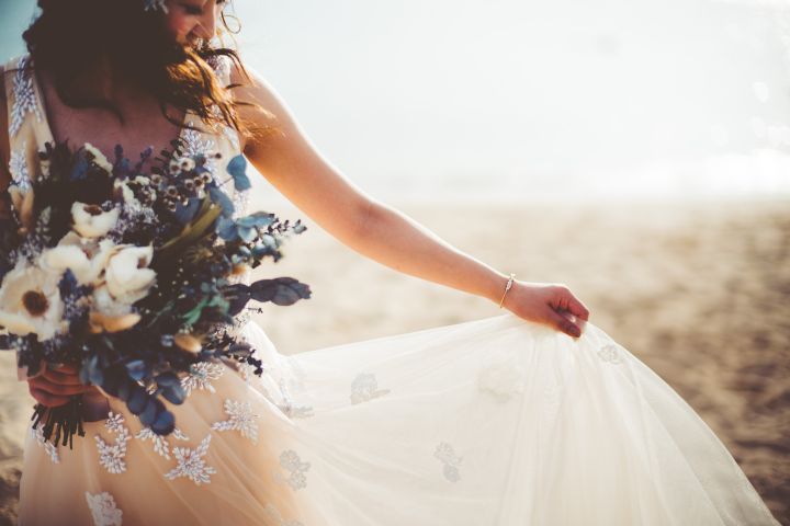 右手にブーケ、左手はドレスの裾をつまんでビーチで風を纏っている花嫁カット
